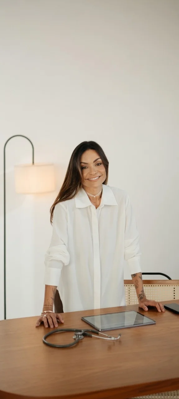 Dra. Claudia Cogo em pé junto à sua mesa de atendimento, sorrindo